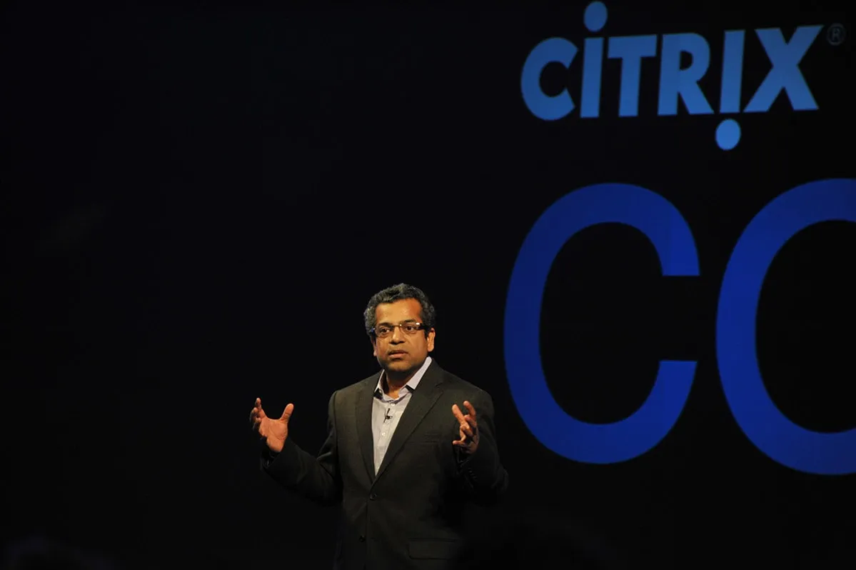 A speaker on stage at a conference, the brand screen behind them