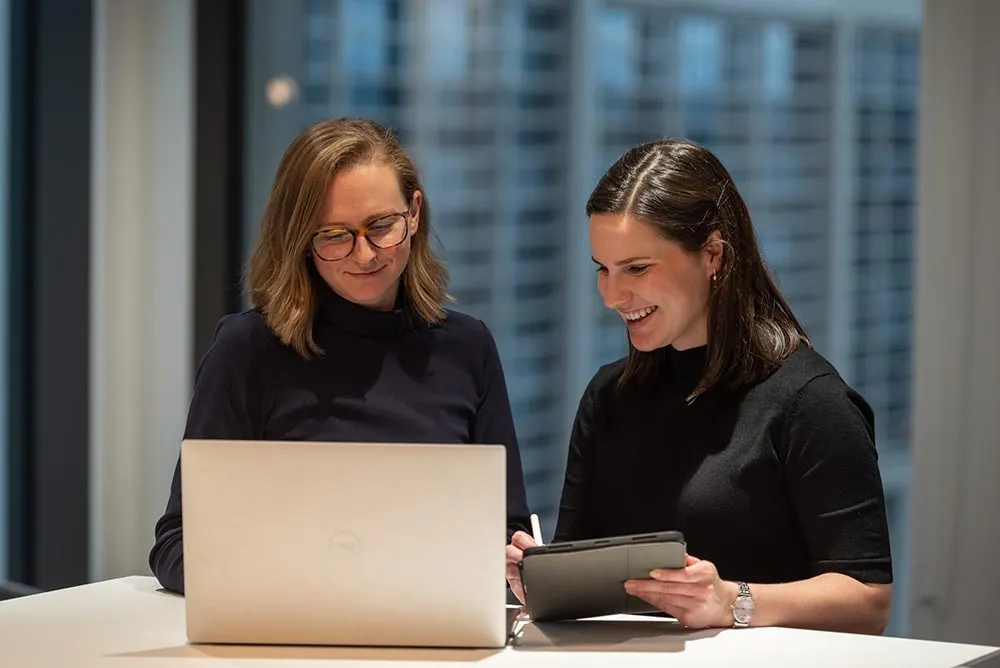 Two colleagues working at a laptop beside a window, city towers behind them