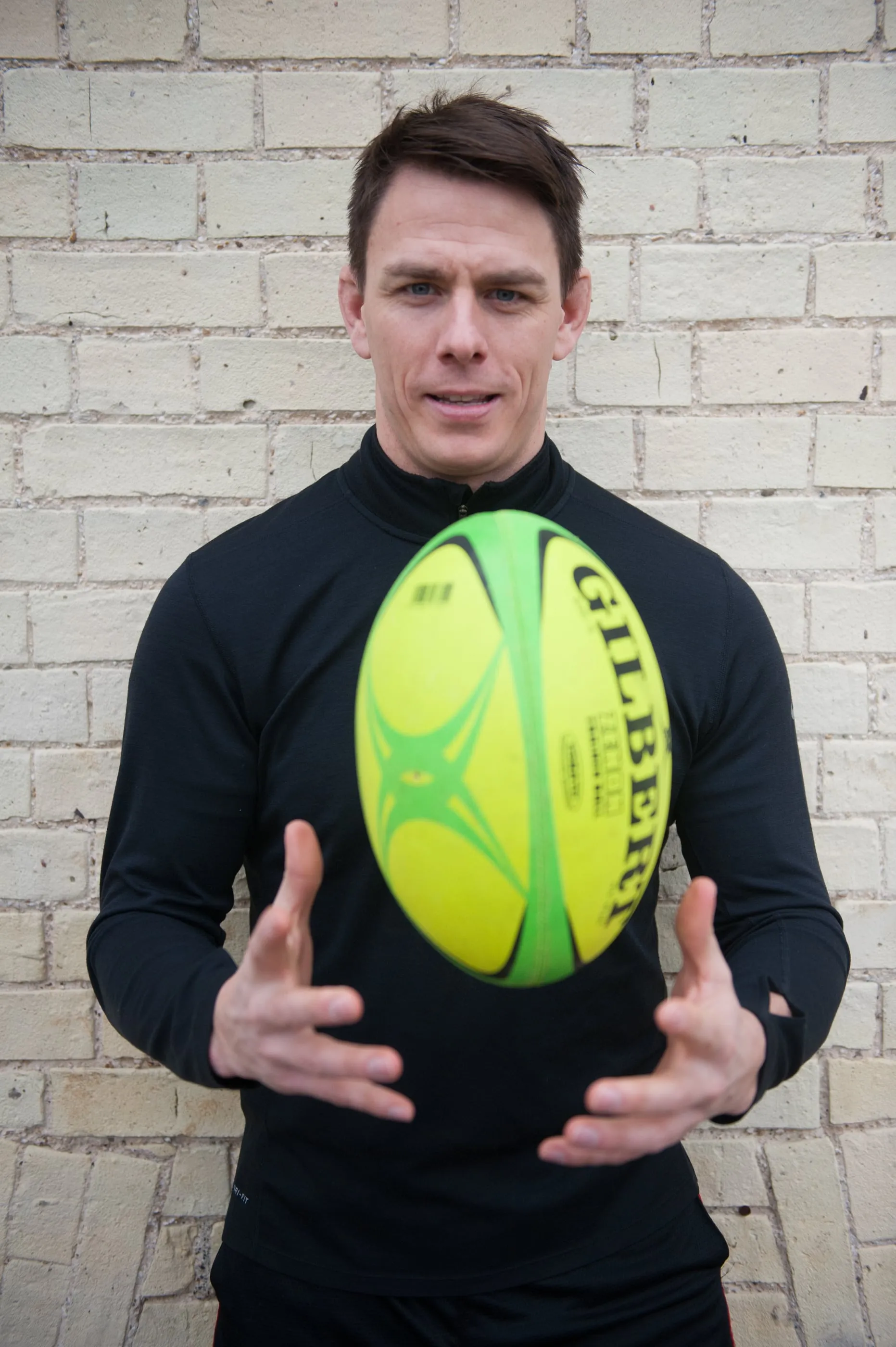A portrait of an athlete holding a rugby ball, photographed against a brick wall